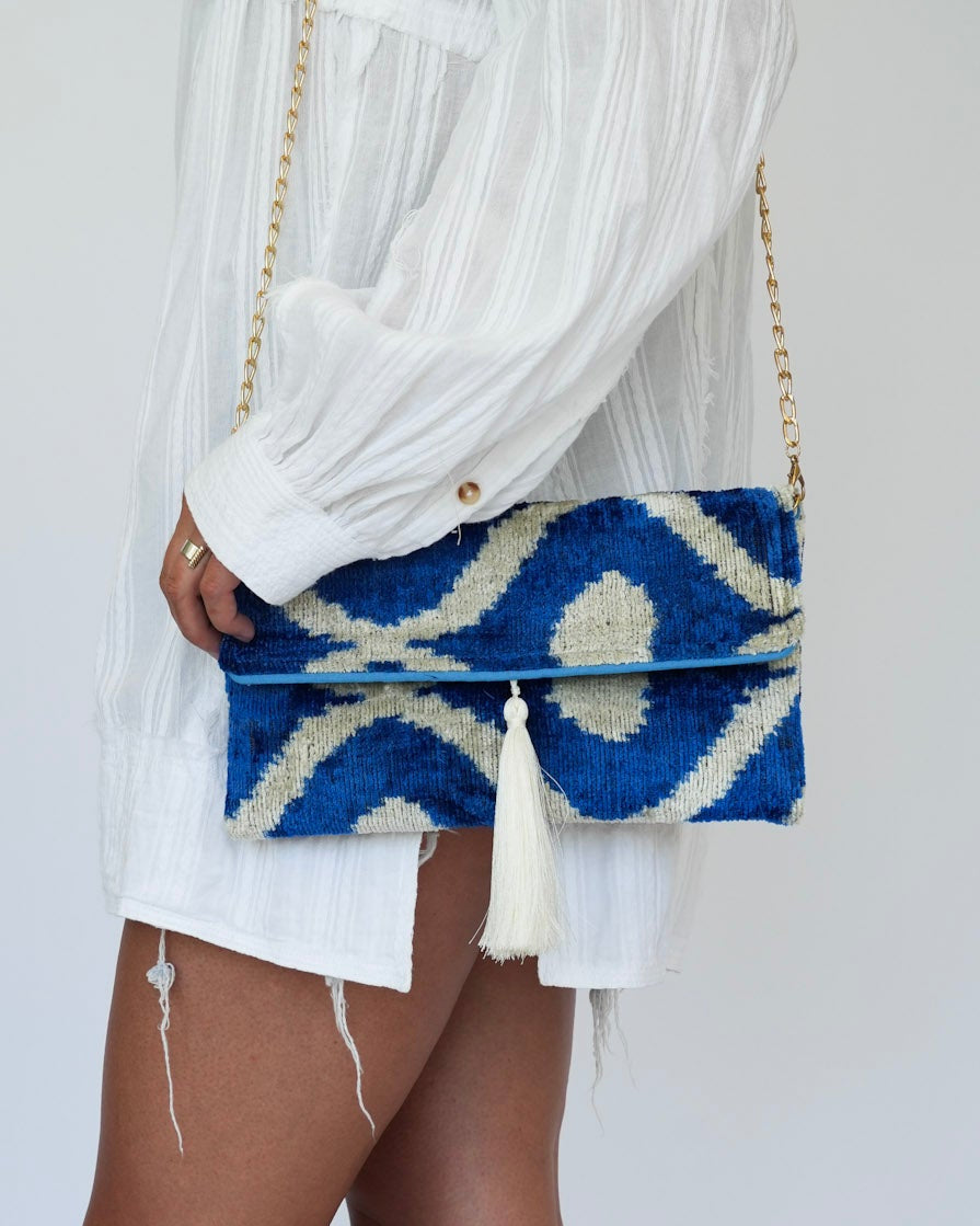 A person wearing a white long-sleeve shirt and shorts, holding a blue and white patterned woven clutch bag with a gold chain strap, paired with beige woven mule sandals against a plain light gray background.