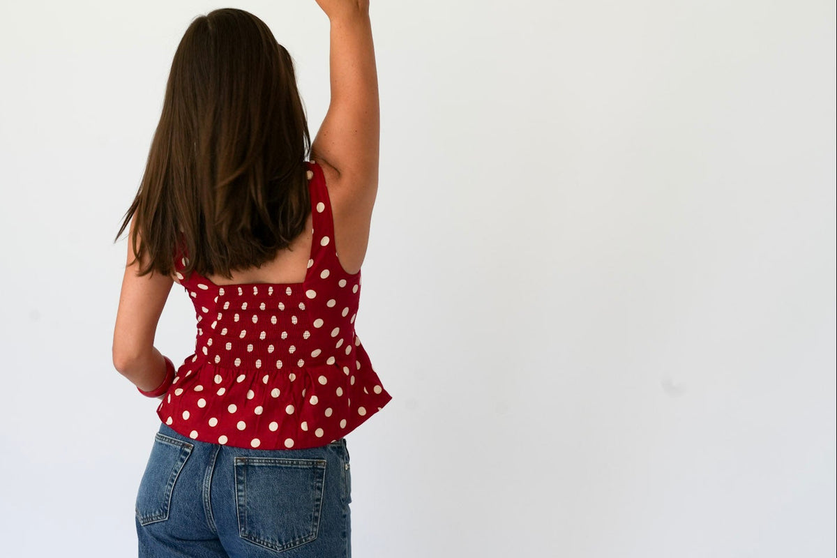 A woman is shown from behind wearing a red sleeveless top with white polka dots and blue jeans, standing against a plain white studio background.