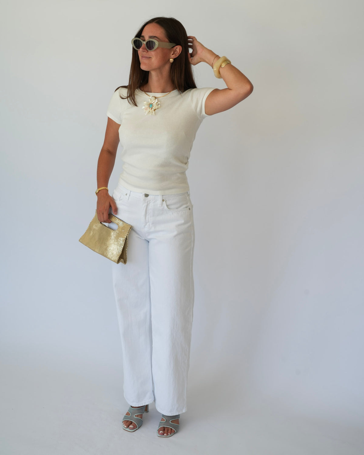 A woman modeling a cream short-sleeve knit top with a gold floral embellishment at the neckline, paired with white wide-leg jeans, a gold clutch, and silver sandals against a plain light gray background.