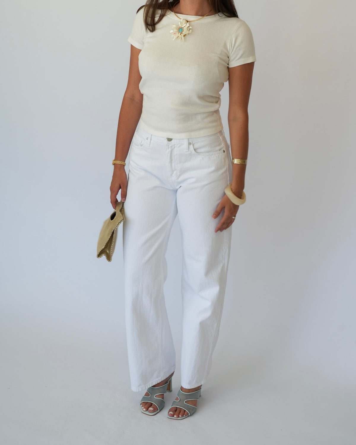 A woman modeling a cream short-sleeve knit top and white wide-leg jeans, accessorized with a gold starburst brooch, gold bracelets, and gray strappy heels, against a plain light gray background.
