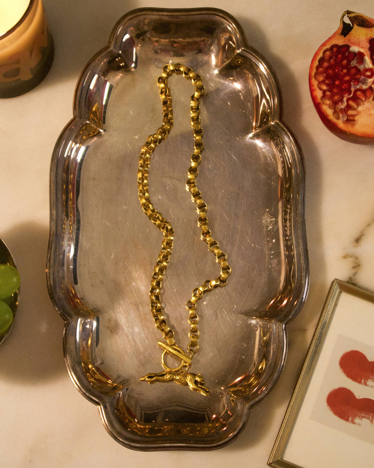 A gold beaded necklace with a toggle clasp is displayed on a silver metal tray, surrounded by lifestyle props including a lit candle, a pomegranate, and a small framed picture on a marble surface.
