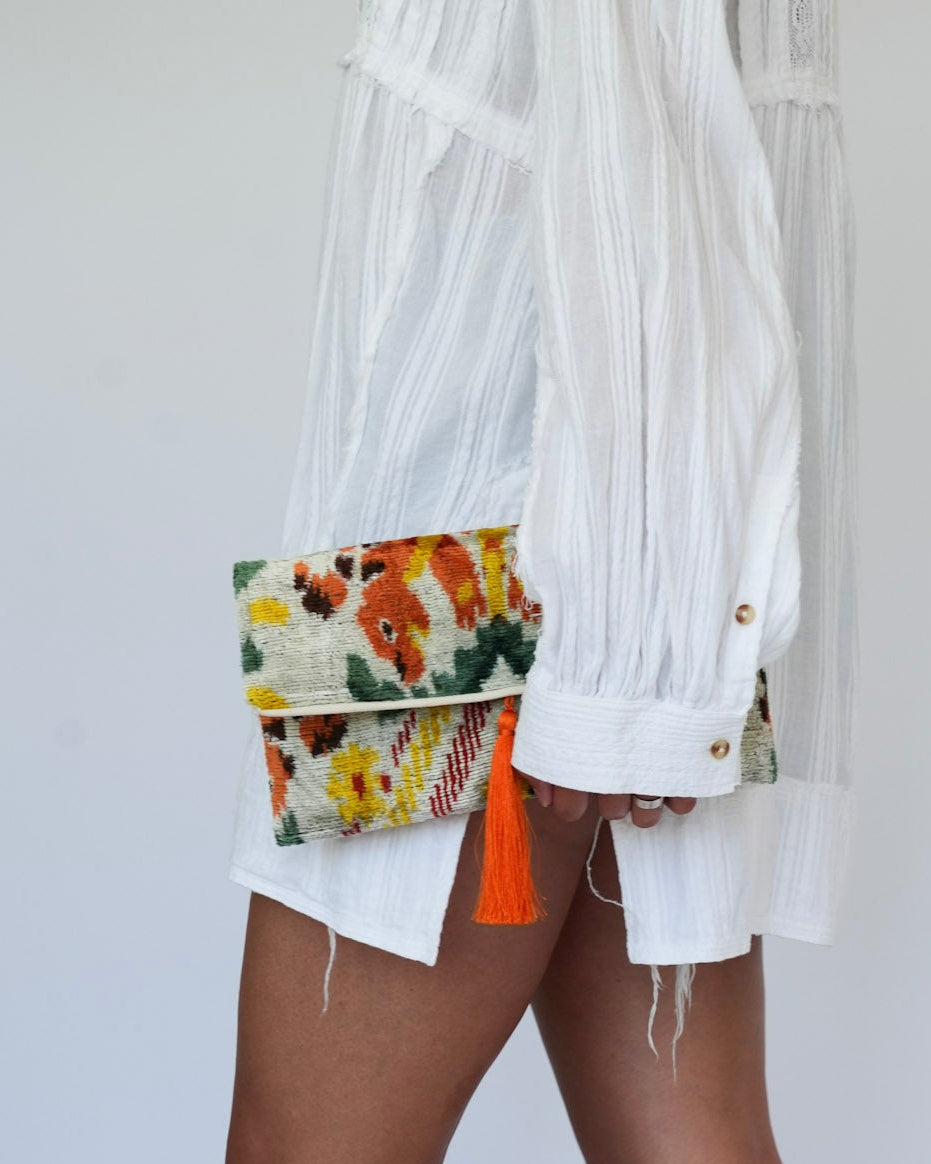 A person wearing a white sheer long-sleeve shirt, white shorts, and beige woven mule sandals, holding a small floral clutch bag with orange tassels against a plain light gray background.